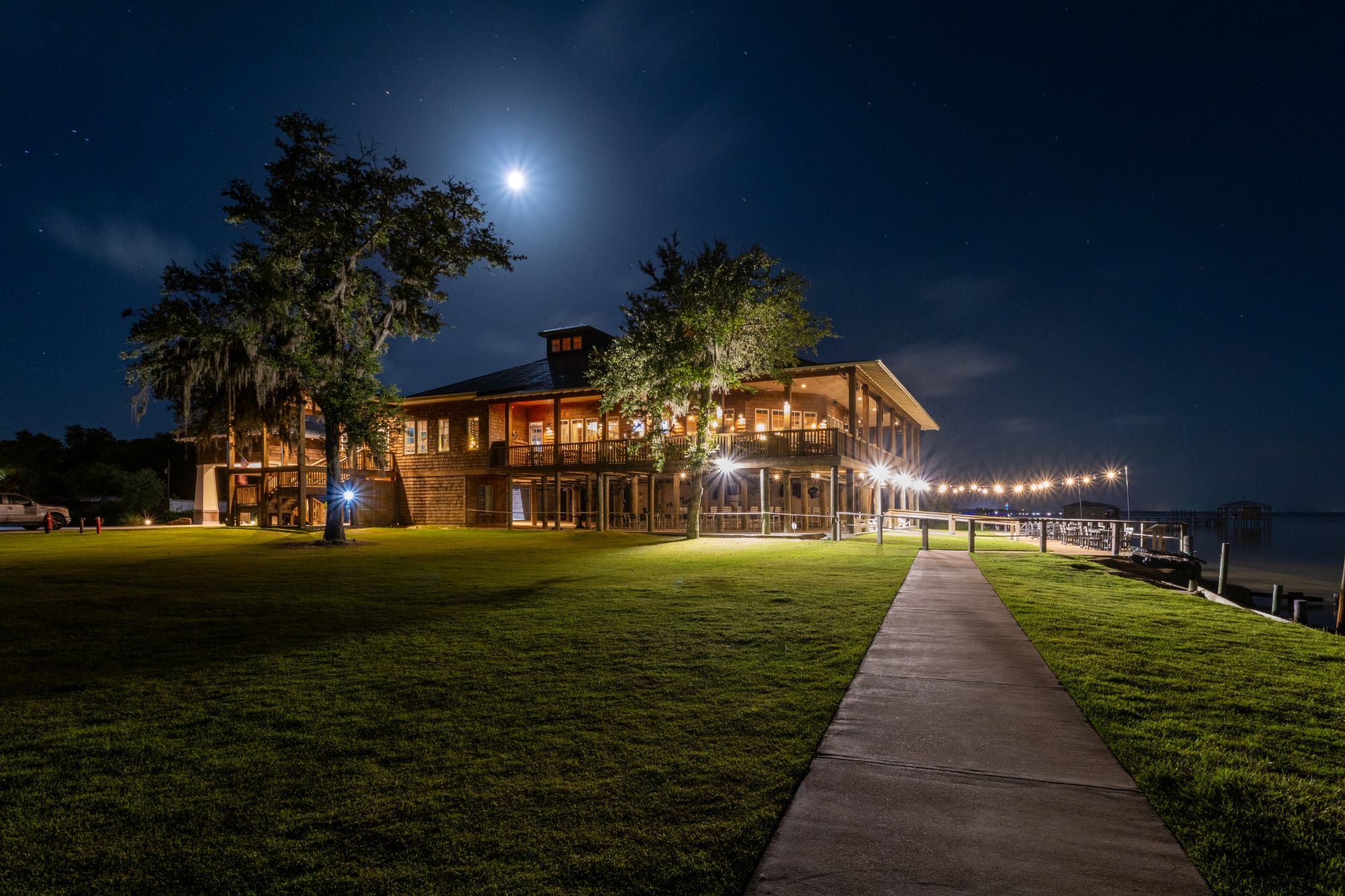 Waterfront event venue lit up at night with string lights and a full moon overhead