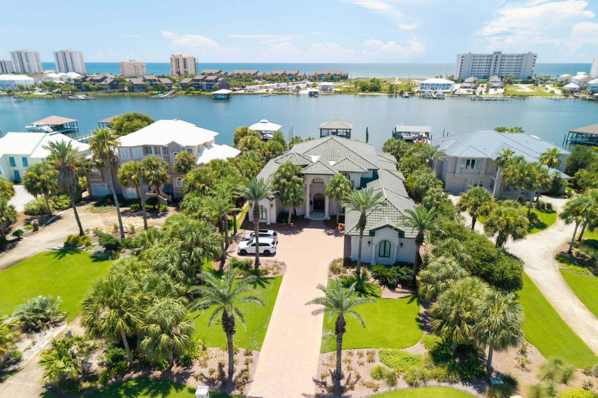Aerial view of a Mediterranean-style waterfront estate with a paver driveway and palms, the bay and Gulf beyond